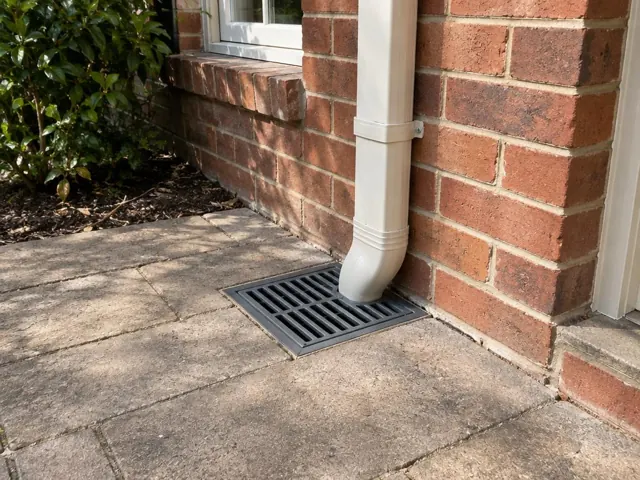 Stormwater grate beside a brick house wall and downpipe on a dry paved patio in Gladesville